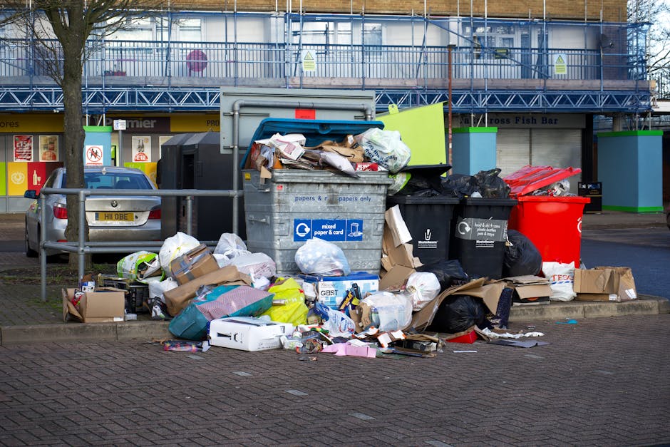A large collection of mixed household waste and rubbish accumulated in an outdoor public area, featuring overflowing black, gray, and red waste bins, with various paper, cardboard boxes, plastic bags,