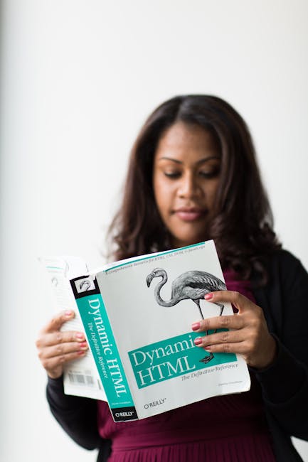 A woman with dark, shoulder-length hair and light brown skin is standing indoors against a plain white background, holding and reading an open book titled 'Dynamic HTML.' She is dressed in a black bla